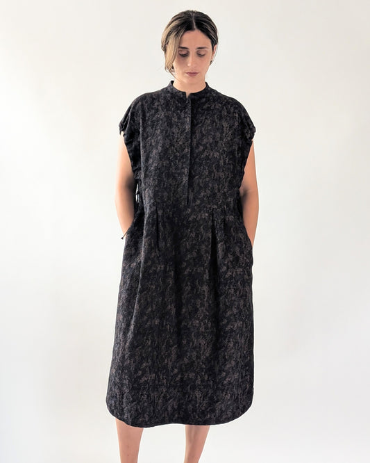 A woman wearing a dark navy textured midi dress with cap sleeves, a high collar, and side pockets, standing against a plain white studio background.