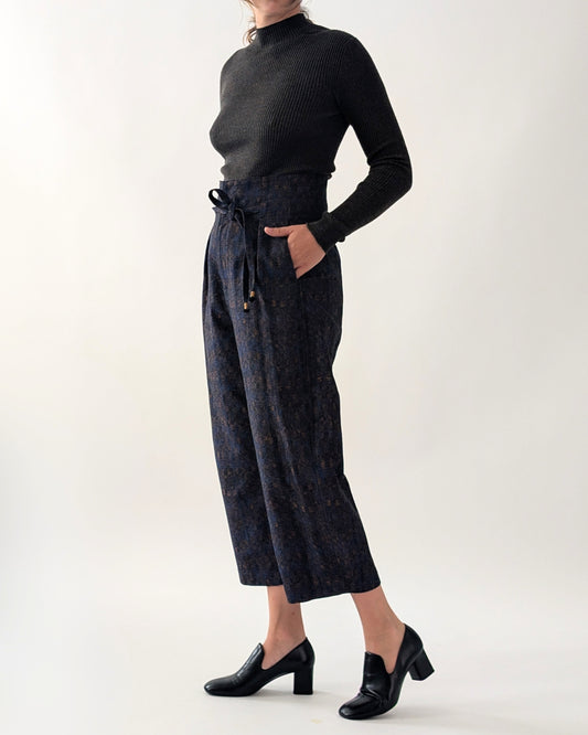 A model wearing a black ribbed mock-neck long-sleeve top and dark navy patterned cropped trousers with a drawstring waist, paired with black block-heel loafers, against a plain light grey studio background.