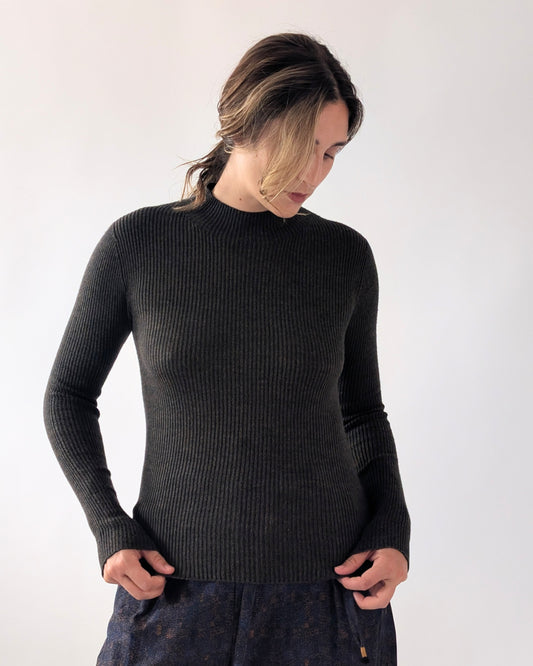 A woman wearing a black ribbed knit mock neck sweater, paired with dark patterned trousers, standing against a plain light grey background.