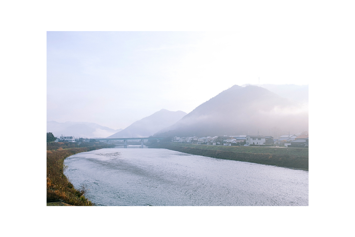 A scenic landscape photograph showing a wide river flowing through a valley, with misty mountains in the background and a bridge spanning the water.