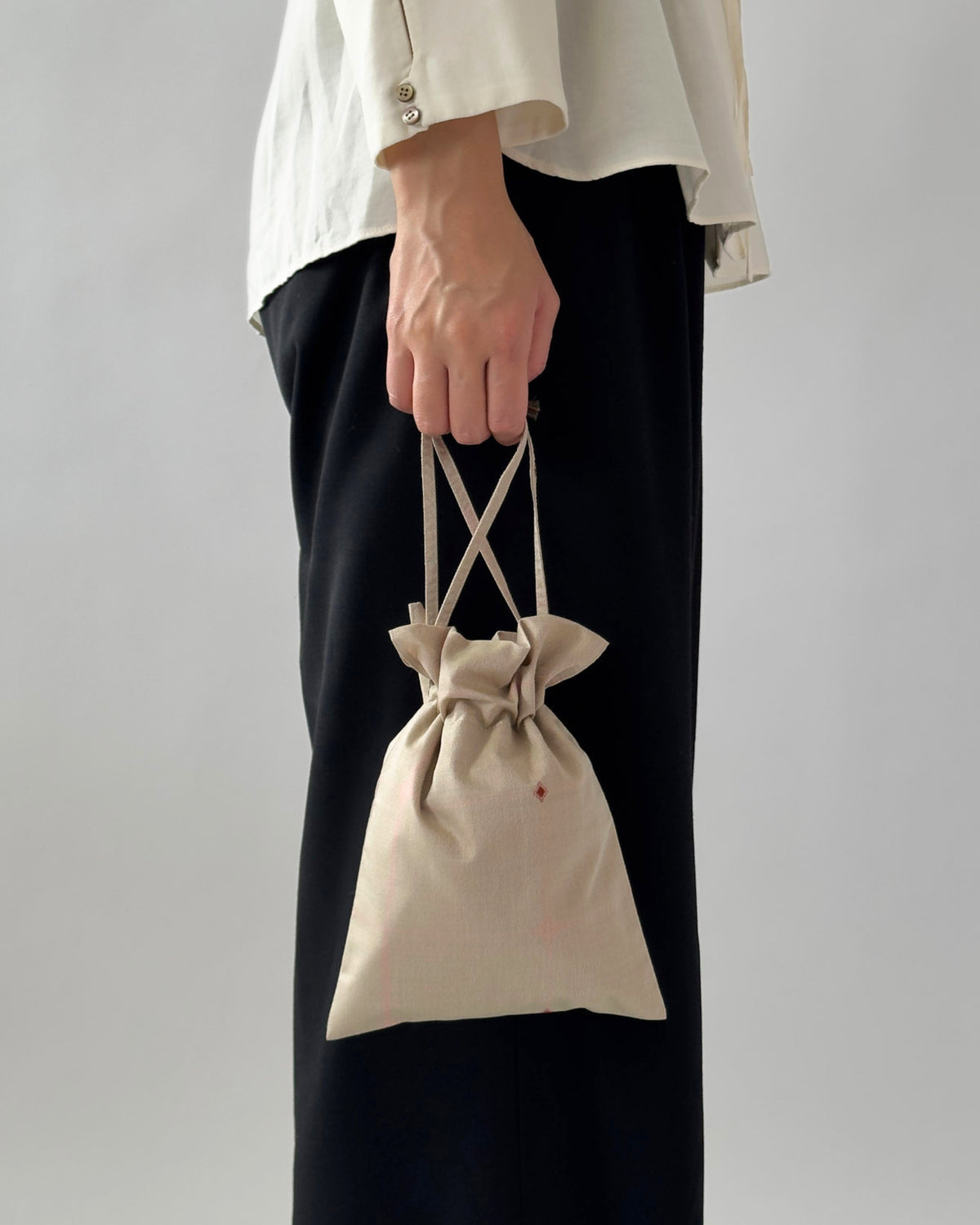 A person wearing a white blouse and black wide-leg pants holds a small beige drawstring pouch bag with crisscross top handles against a plain light gray background.