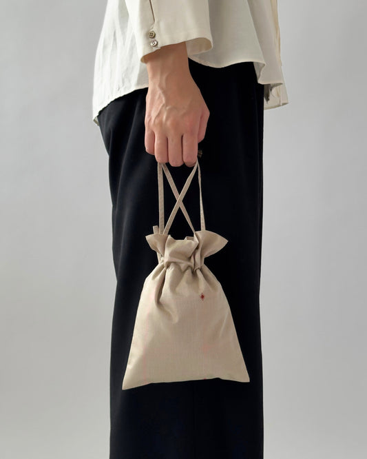 A person wearing a white blouse and black wide-leg pants holds a small beige drawstring pouch bag with crisscross top handles against a plain light gray background.