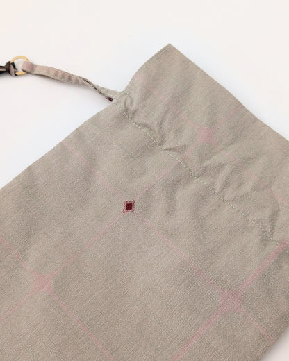 A close-up of a light beige fabric bag or pouch with a subtle pink grid pattern, featuring a drawstring closure and a small dark red stitched accent.