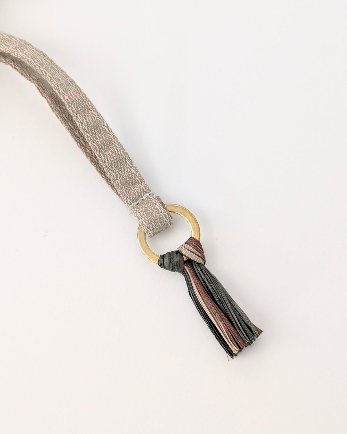 A close-up of a woven strap in a light beige tone with a gold-tone ring and a tassel featuring multiple colors including black, brown, and pink, set against a plain white background.