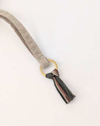 A close-up of a woven strap in a light beige tone with a gold-tone ring and a tassel featuring multiple colors including black, brown, and pink, set against a plain white background.