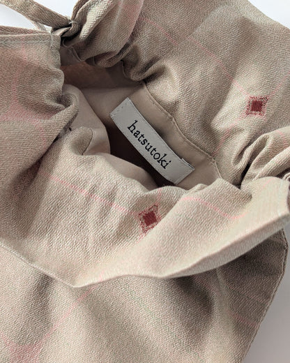 Close-up of a beige fabric bag or pouch with a drawstring closure and a visible white brand label reading hatsutoki. The fabric has a soft woven texture with faint pink lines and small muted red square motifs.