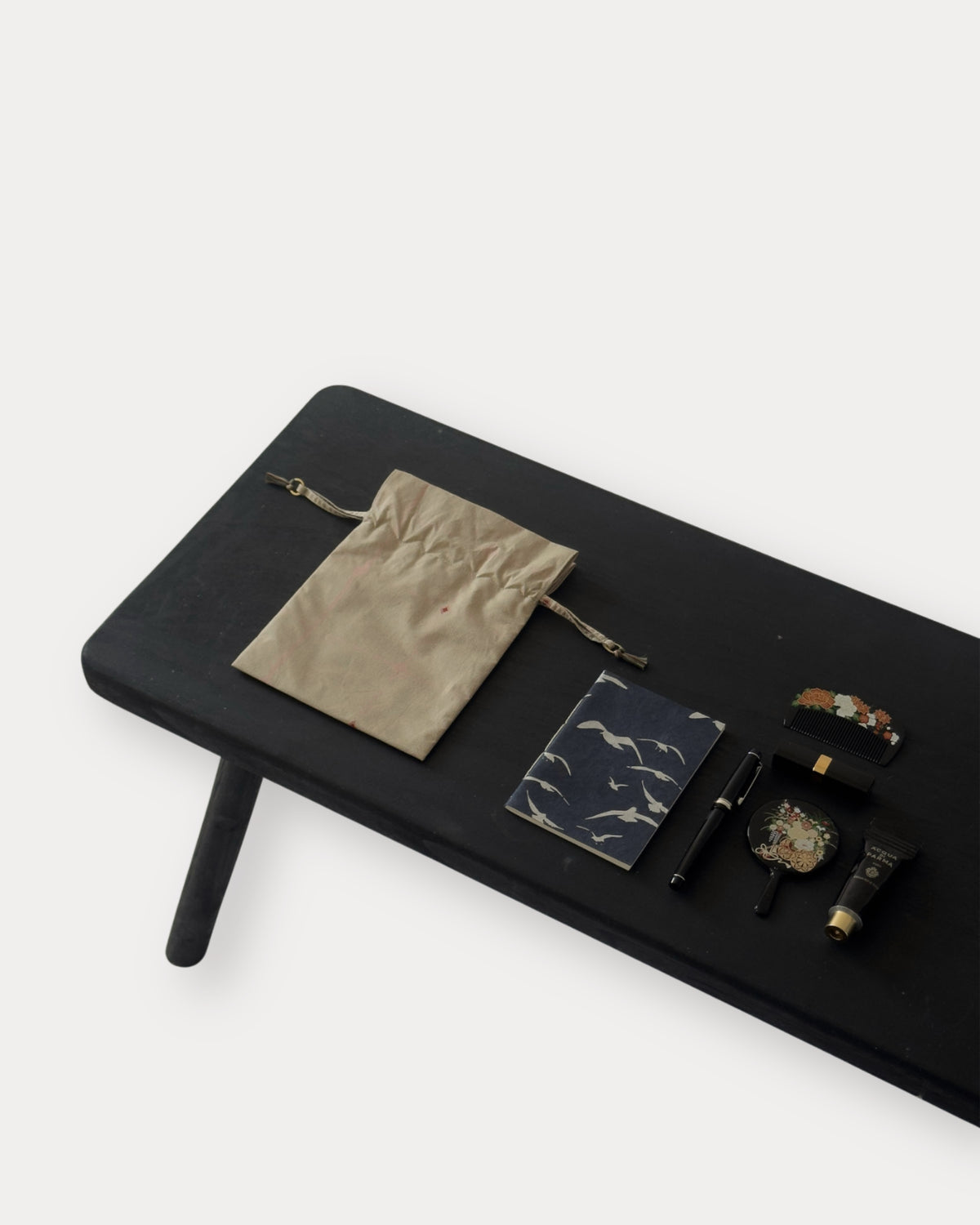 A styled flat-lay of various small accessories and cosmetics arranged on a black rectangular table against a light gray background. The items include a beige drawstring pouch, a dark marble notebook, a black pen, a watch, a tube of cream, and a small box.