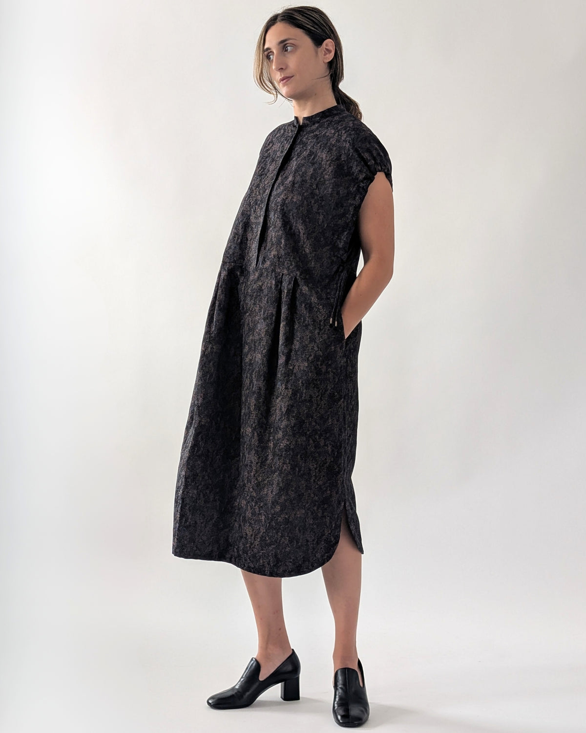 A woman modeling a dark navy sleeveless midi dress with a textured, mottled pattern, standing against a plain light gray studio background.