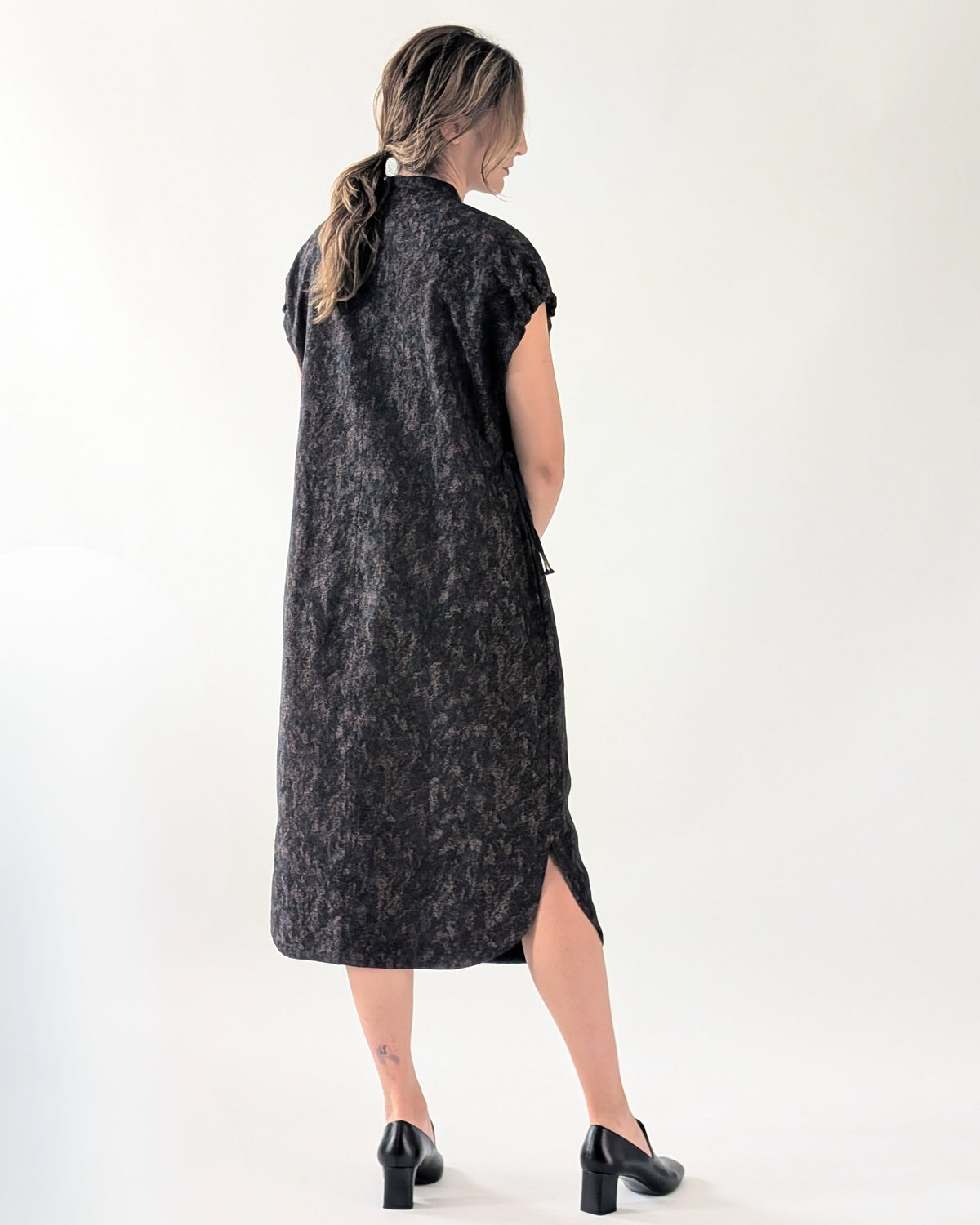 A woman is shown from behind wearing a sleeveless, knee-length dark navy dress with a subtle textured pattern and a side slit, paired with black heels, against a plain white studio background.