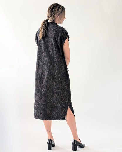 A woman is shown from behind wearing a sleeveless, knee-length dark navy dress with a subtle textured pattern and a side slit, paired with black heels, against a plain white studio background.