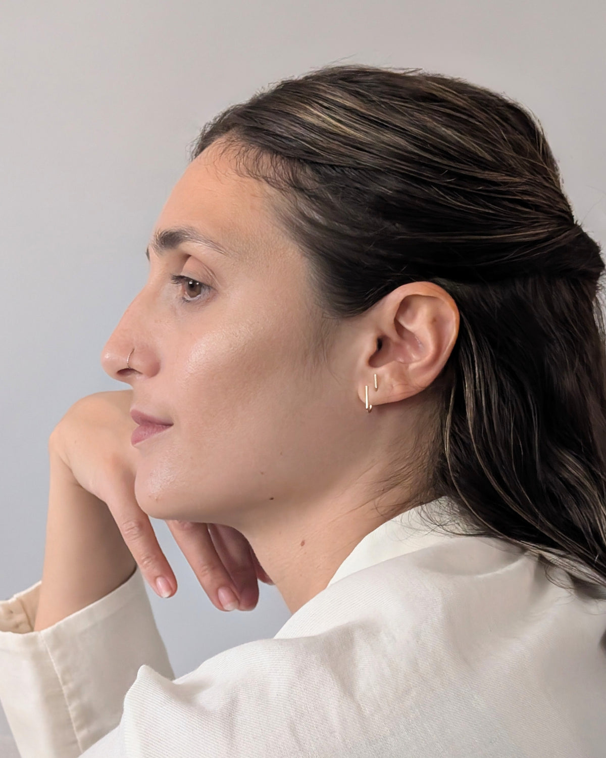 A woman in profile wearing a delicate 14KGF gold hoop earring, resting her chin on her hand against a plain light gray background.