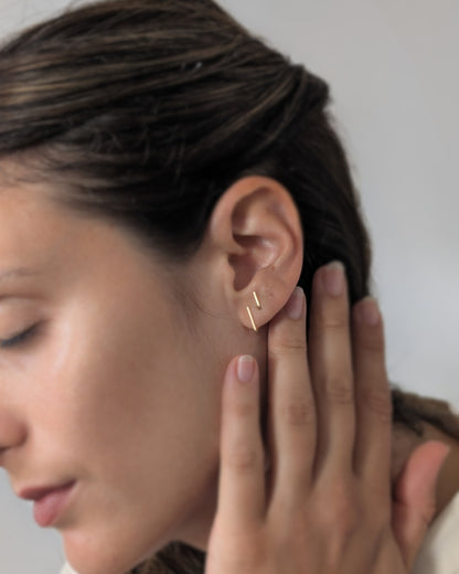 A close-up of a woman's ear wearing a delicate 14KGF gold ear cuff with two parallel diagonal bars, with her hand resting near her face against a plain light background.