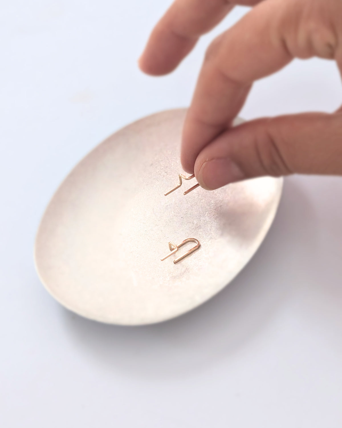 A close-up of a hand placing two small 14KGF gold-toned earrings onto a round, metallic gold tray against a plain white background.