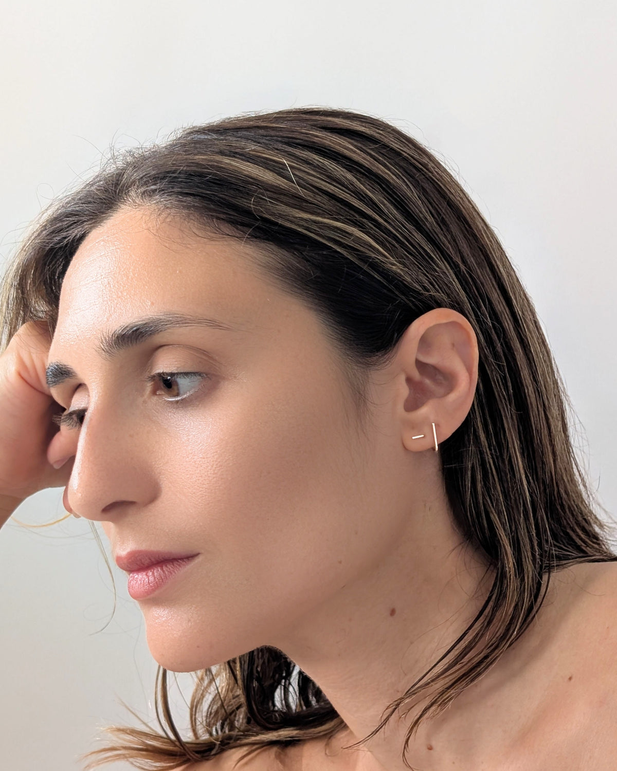 A close-up side profile of a woman with dark brown hair wearing a delicate 14KGF gold geometric stud earring on her earlobe, set against a plain light grey background.