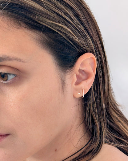 A close-up of a woman's ear wearing a delicate 14KGF gold-toned earring with a thin vertical bar, shown against a light neutral background.