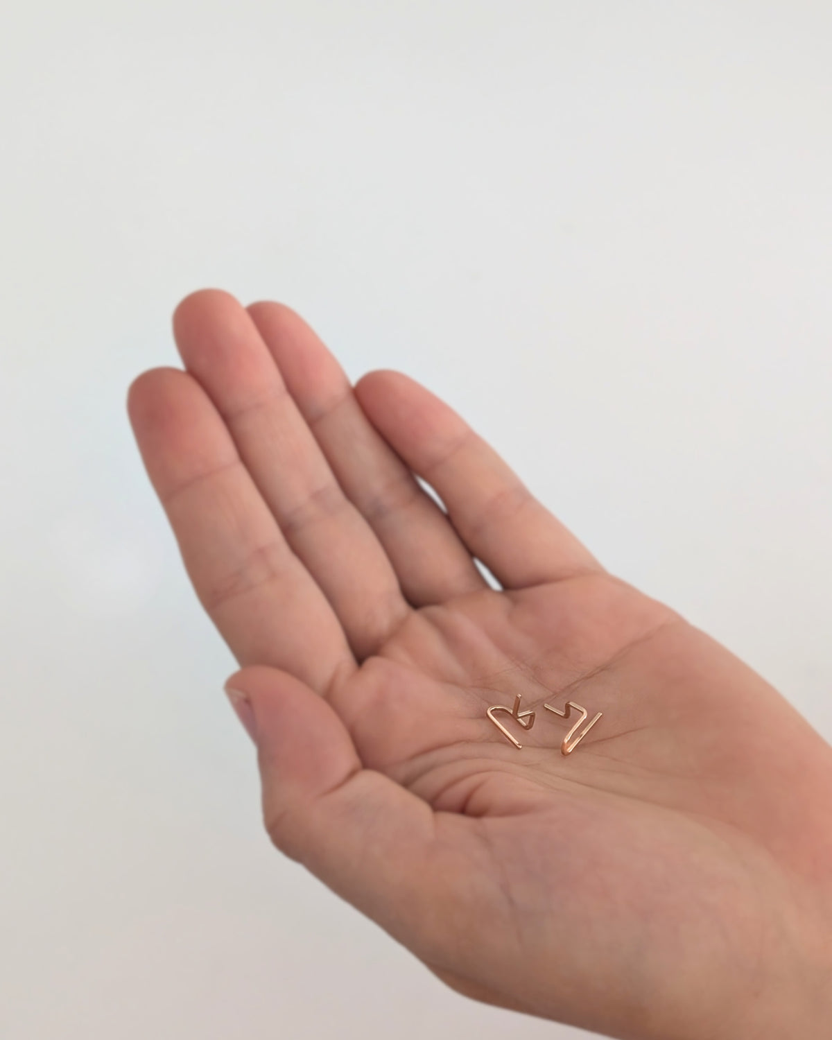 A close-up of a person's open palm holding a pair of tiny 14KGF gold-toned geometric stud earrings against a plain light background.