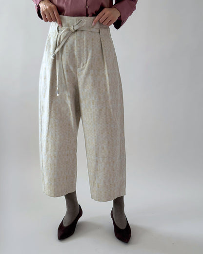 A person wearing high-waisted cropped wide-leg trousers with a tie waist, in a light beige and white patterned fabric, paired with a dusty rose top, grey socks, and dark burgundy flats against a plain light grey studio background.