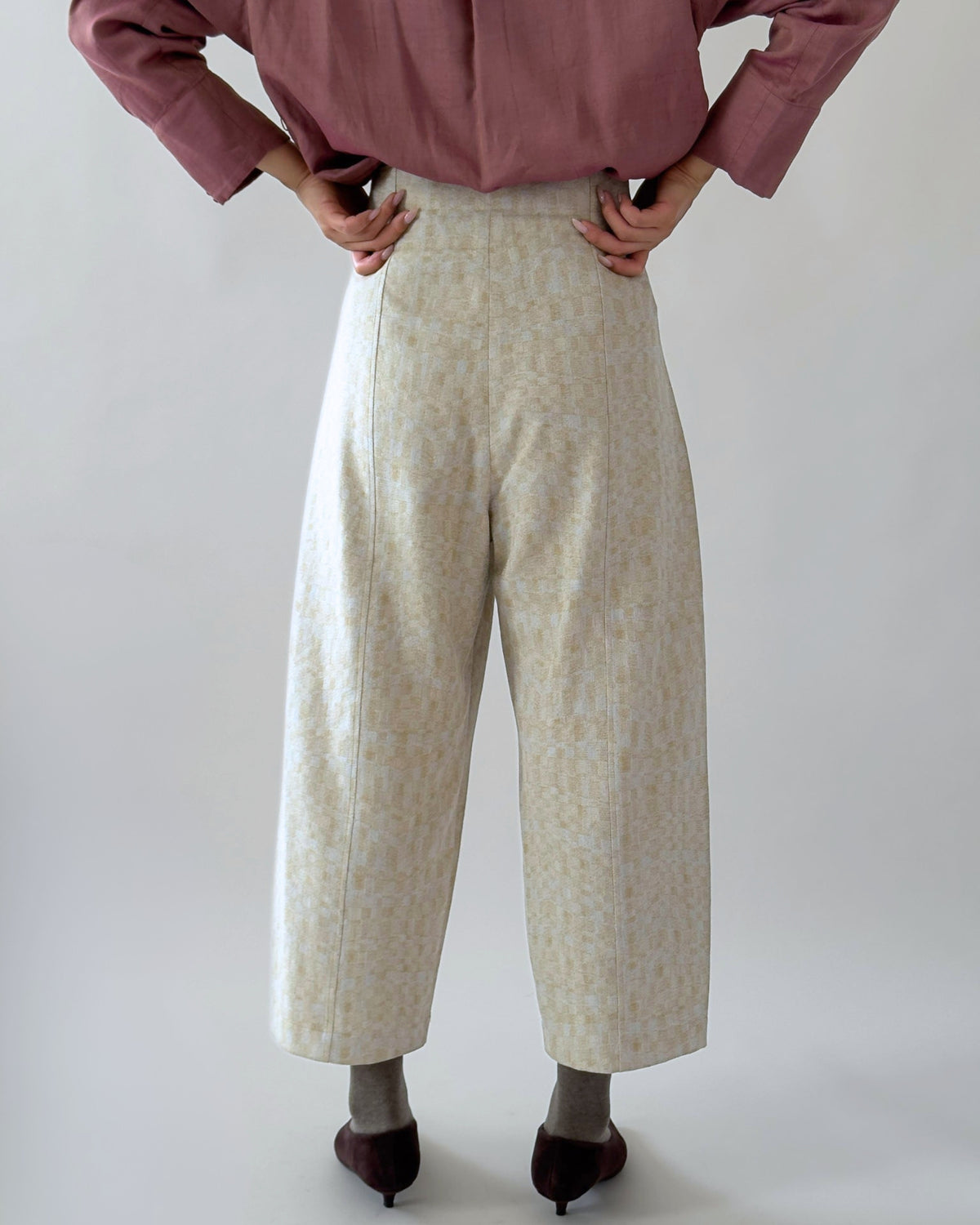 A person shown from the waist down wearing high-waisted, wide-leg cropped trousers in a light beige and white textured pattern, paired with a dusty rose blouse and dark brown pointed-toe flats, against a plain light grey background.