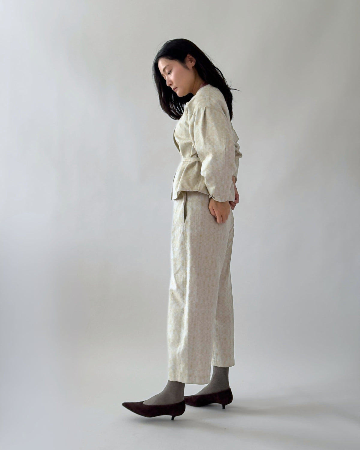 A woman stands in profile wearing a light beige textured two-piece outfit consisting of a long-sleeve top and wide-leg trousers, paired with dark brown pointed-toe heels against a plain light gray studio background.
