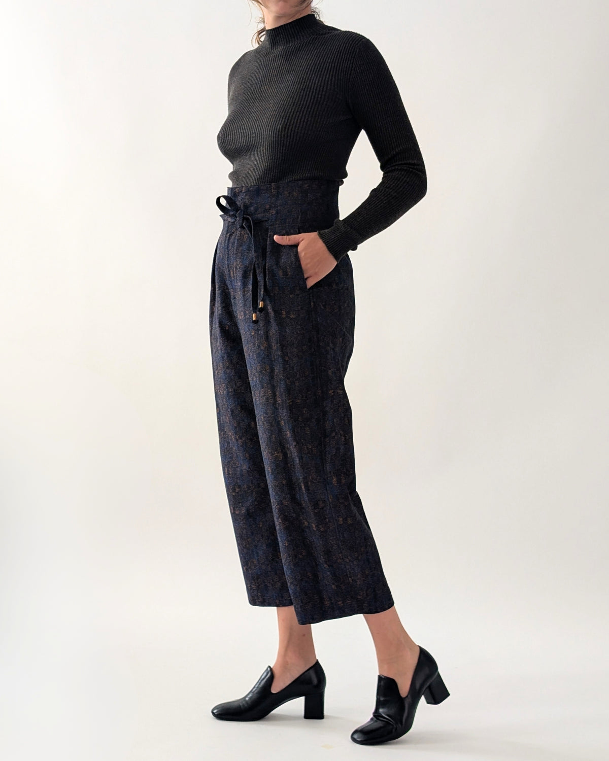 A model wearing a black ribbed mock-neck long-sleeve top and dark navy patterned cropped trousers with a drawstring waist, paired with black block-heel loafers, against a plain light grey studio background.