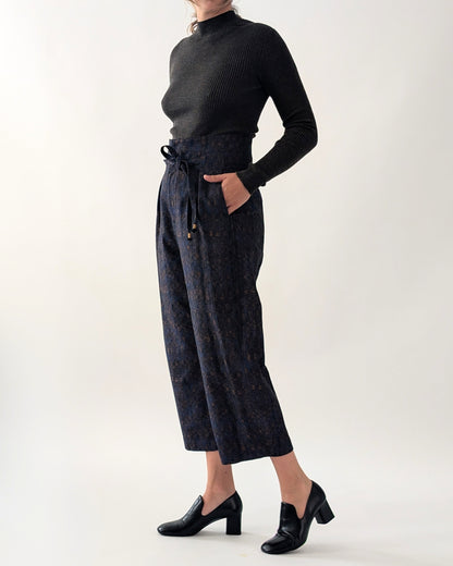 A model wearing a black ribbed mock-neck long-sleeve top and dark navy patterned cropped trousers with a drawstring waist, paired with black block-heel loafers, against a plain light grey studio background.