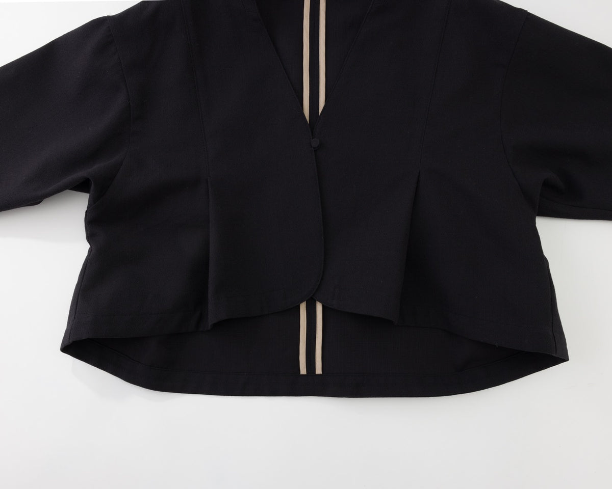 A black long-sleeve cropped cardigan or jacket with a V-neckline, featuring two vertical beige stripes down the front center and a button closure at the collar, laid flat on a white background.