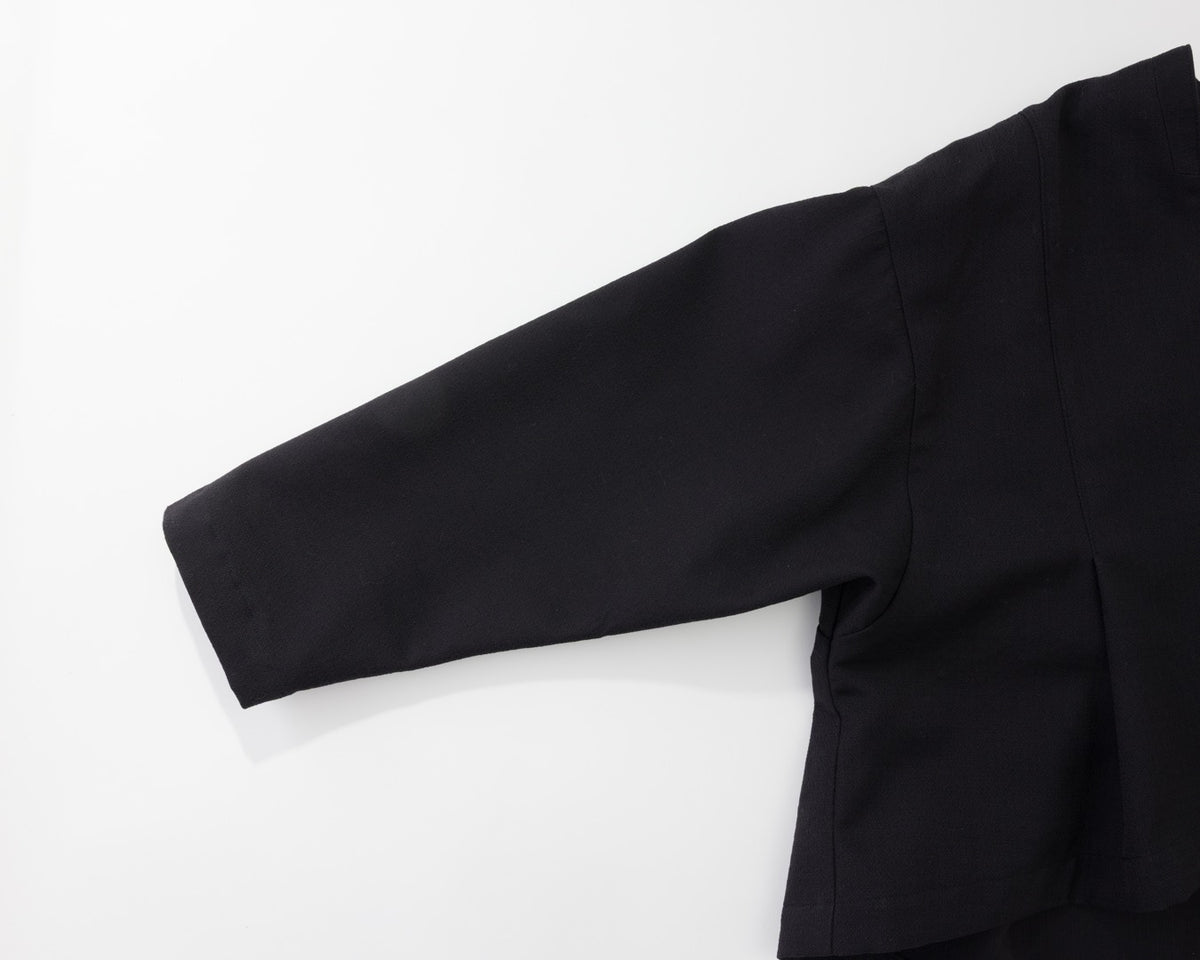 A close-up of a black long-sleeve garment laid flat on a white background, showing the sleeve, shoulder seam, and side drape.