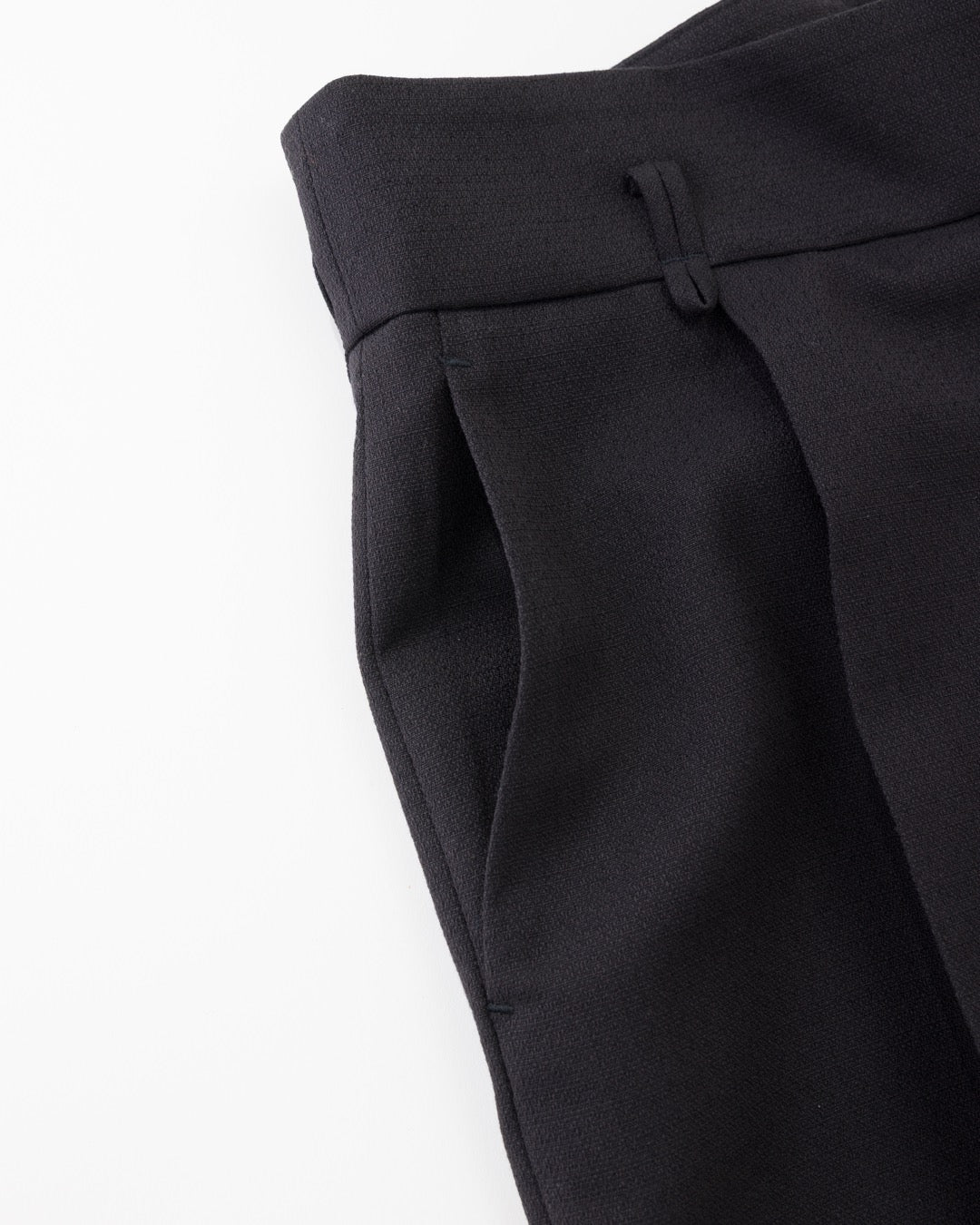 Close-up of black tailored trousers showing the waistband, front pleat, and side pocket on a plain white background.