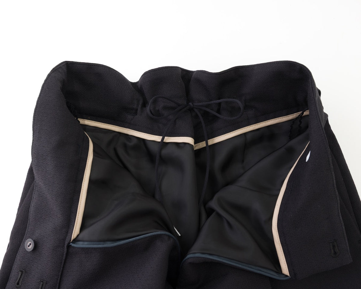 Close-up of a black tailored jacket or coat laid flat on a white background, showing the inner lining, drawstring waistband, and contrast light beige piping.