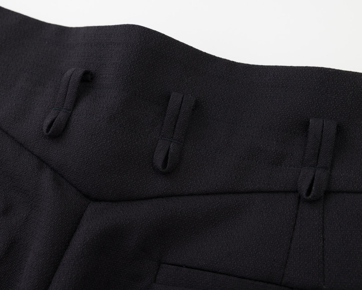 Close-up of a black textured fabric garment, showing pleated details and a pocket on a white background.