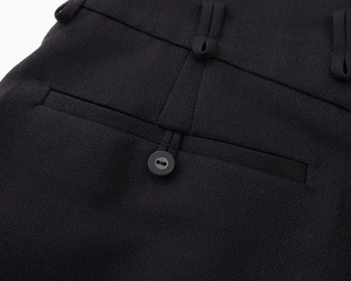 Close-up of black tailored trousers showing the waistband, belt loops, front pleat, and a buttoned welt pocket.