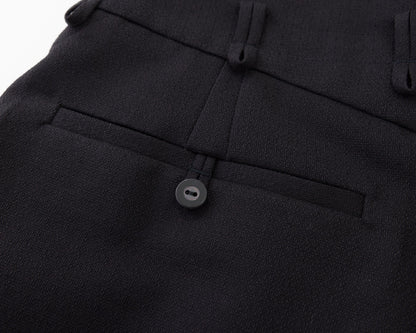 Close-up of black tailored trousers showing the waistband, belt loops, front pleat, and a buttoned welt pocket.