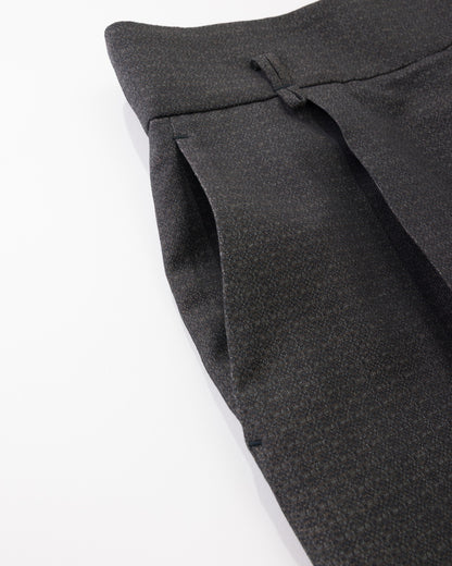 Close-up of black pleated trousers showing the fabric texture and waistband detail against a white background.
