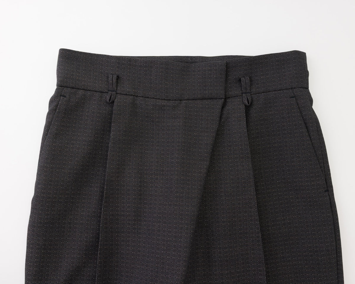 Close-up of the waistband and upper front of black pleated trousers, showing belt loops, front fly, and side pockets on a plain light background.