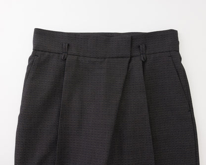 Close-up of the waistband and upper front of black pleated trousers, showing belt loops, front fly, and side pockets on a plain light background.
