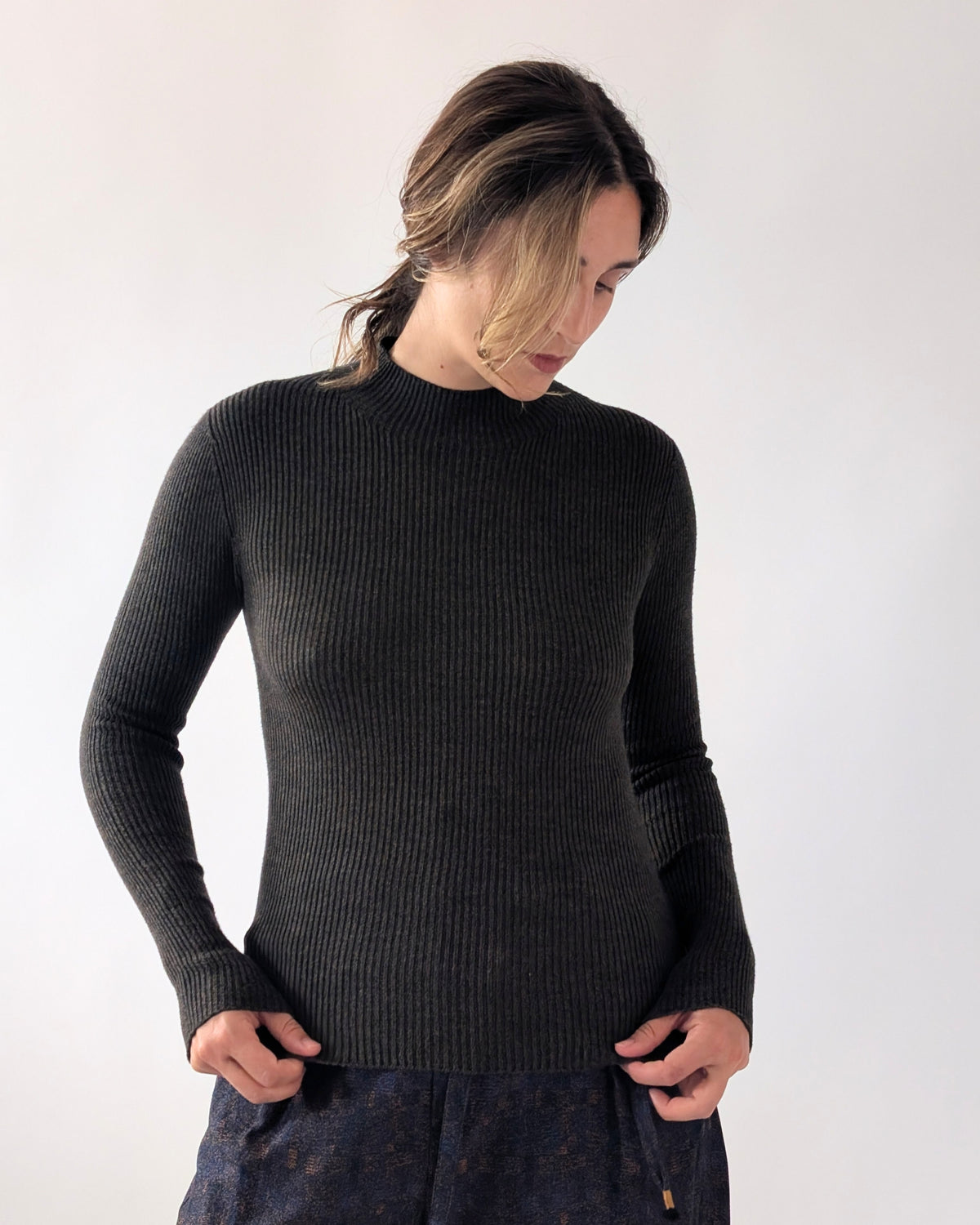 A woman wearing a black ribbed knit mock neck sweater, paired with dark patterned trousers, standing against a plain light grey background.