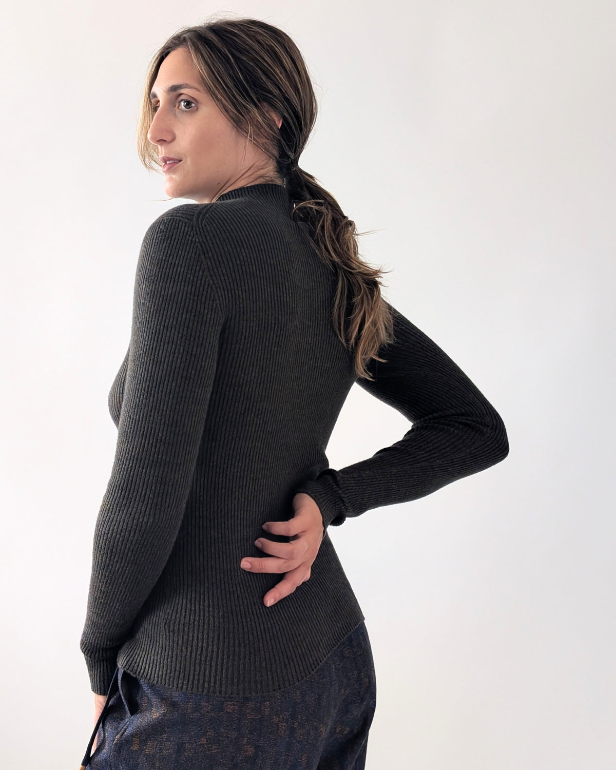 A woman wearing a black ribbed knit sweater, shown from a side-back angle against a plain white studio background.
