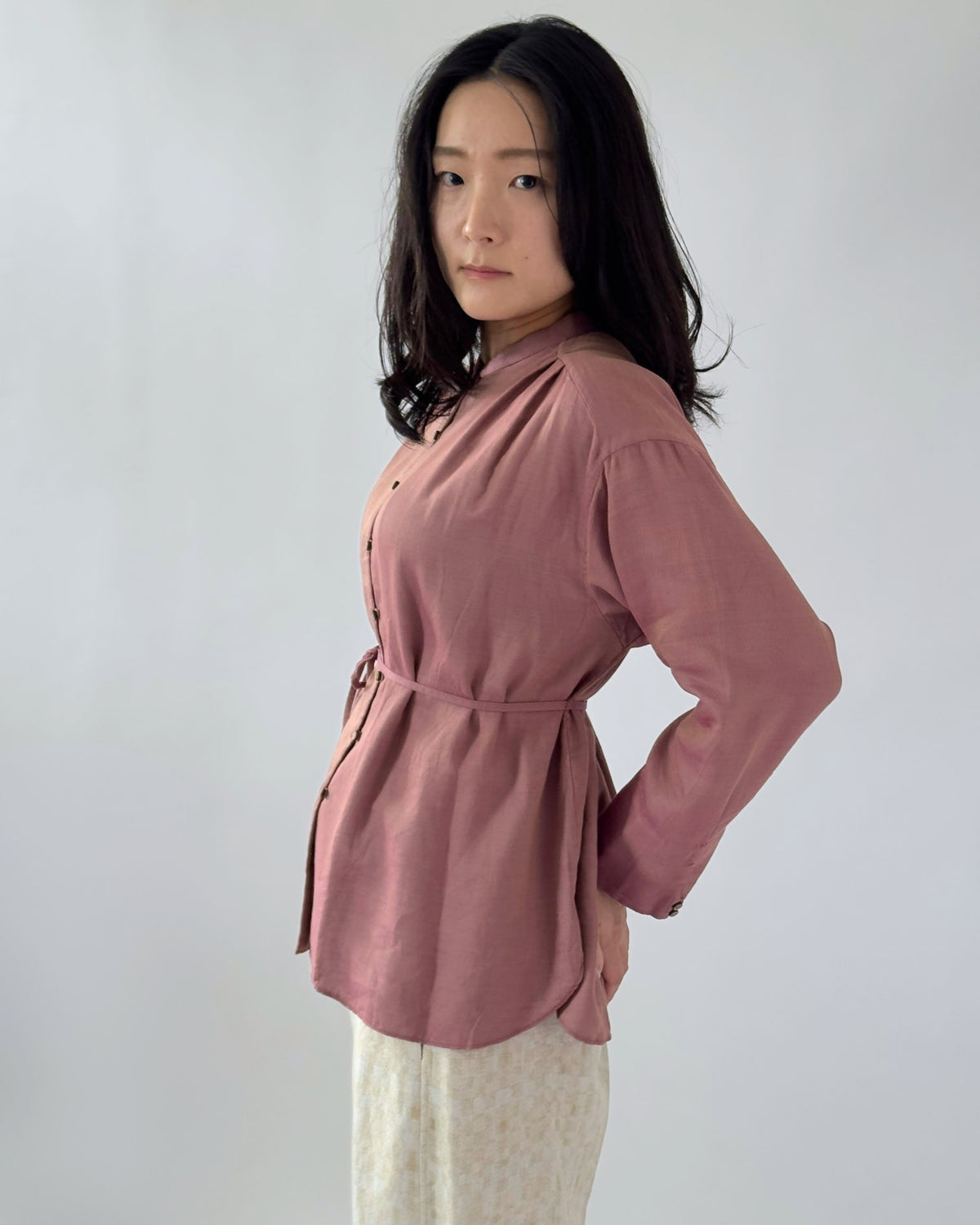 A woman wearing a dusty rose long-sleeve button-up blouse with a tie waist belt, paired with light beige patterned trousers, standing against a plain light grey background.