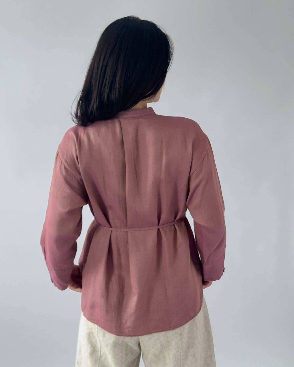 A woman viewed from behind wearing a dusty rose long-sleeve wrap-style blouse with a defined waist seam, paired with light beige pants, against a plain light grey background.
