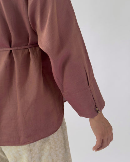 Close-up of a person wearing a dusty rose long-sleeve blouse with a gathered waist and buttoned cuff, paired with light beige patterned bottoms against a plain light grey background.
