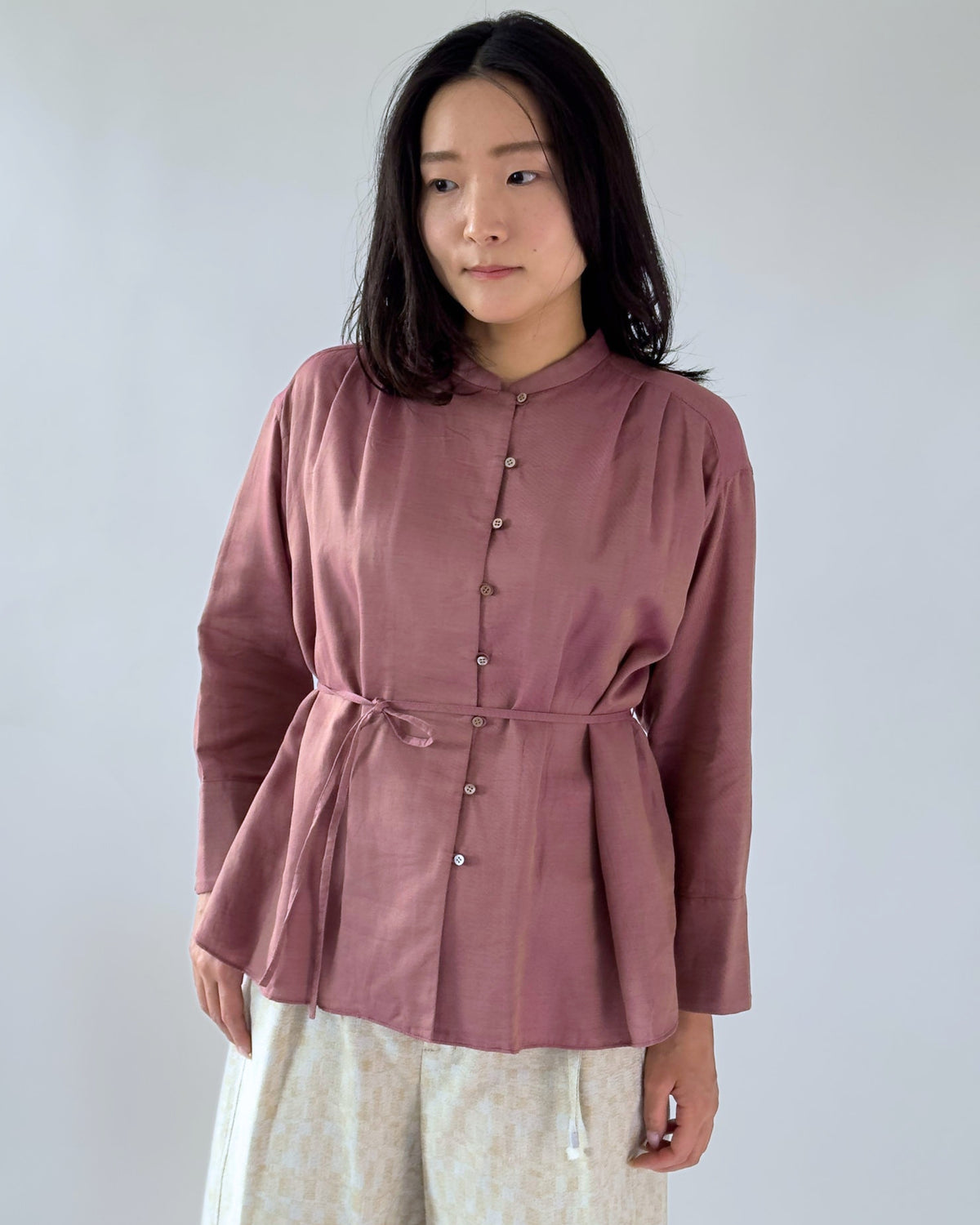 A woman wearing a dusty rose long-sleeve top with a button-down front, stand collar, and matching fabric tie belt at the waist, paired with light beige patterned pants, against a plain light grey background.