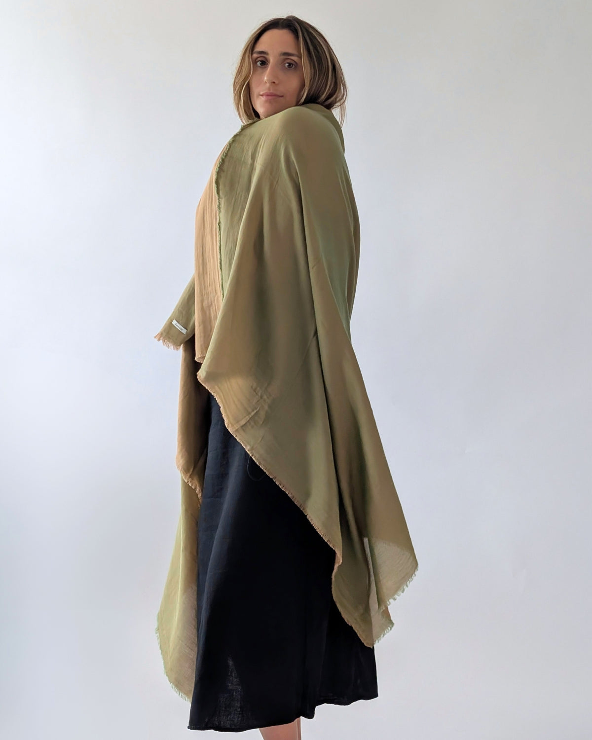 A woman wearing a large green shawl draped over her shoulders, paired with a black midi skirt, photographed against a plain light gray studio background.