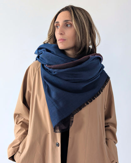 A woman wearing a large navy blue pashmina-style scarf with dark fringe, draped over her shoulders and paired with a tan coat, set against a plain light gray background.