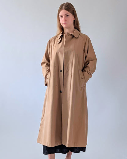 A woman modeling a long, oversized tan trench coat with a collar and front buttons, standing against a plain light gray studio background.