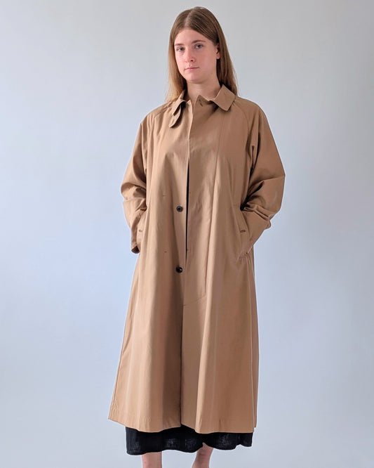 A woman modeling a long, oversized tan trench coat with a collar and front buttons, standing against a plain light gray studio background.