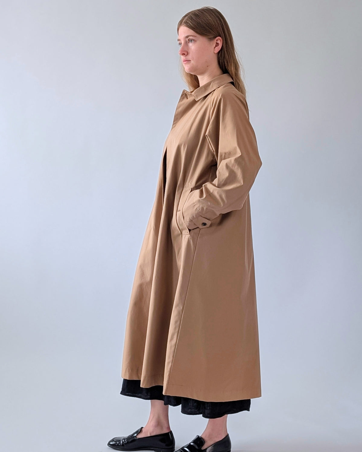A woman modeling a long tan trench coat with a collar and front pockets, paired with black pants and black loafers, standing against a light gray studio background.
