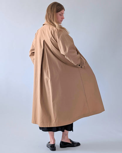 A woman wearing a long camel trench coat, shown from a side-back angle against a light gray studio background.