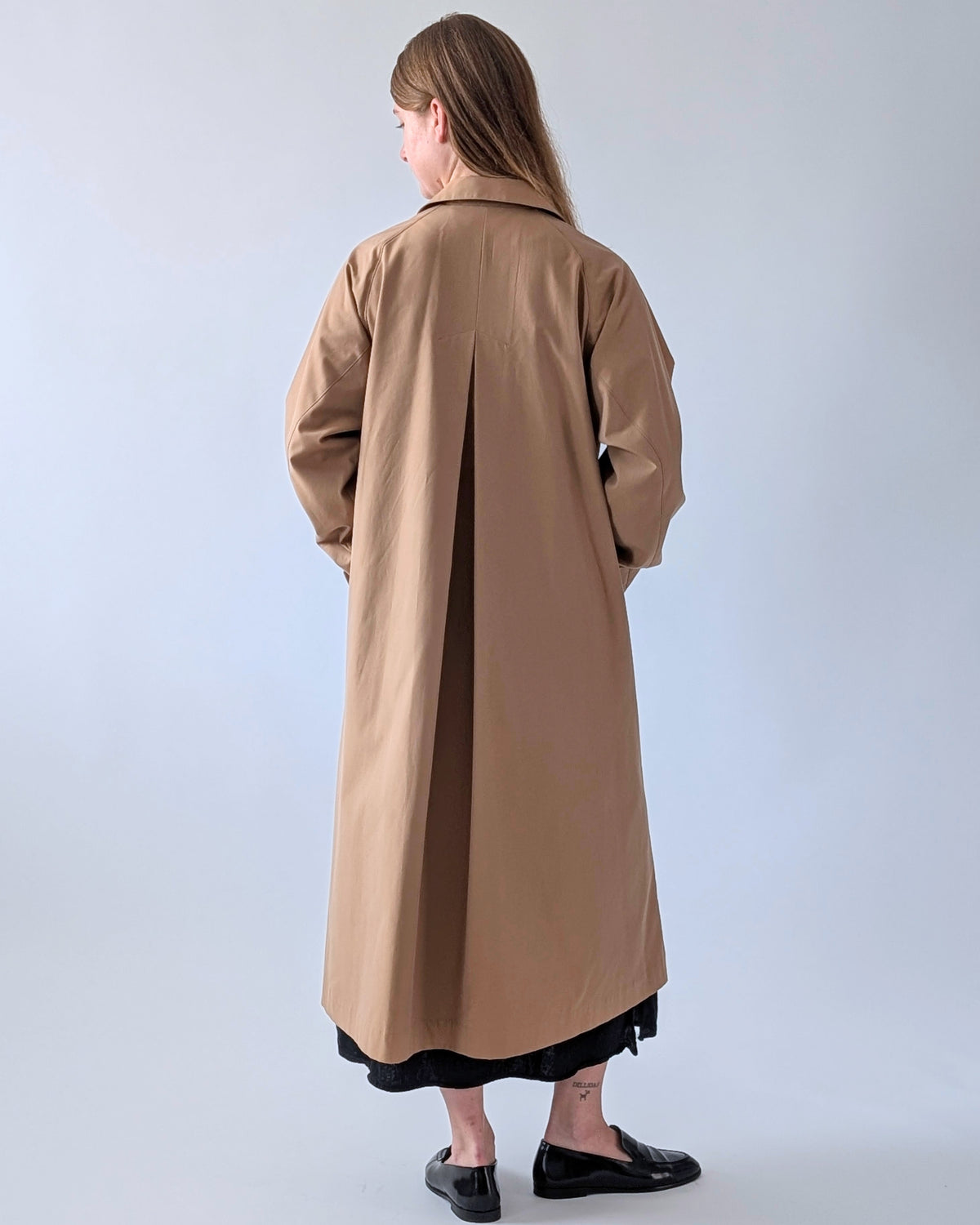A woman is shown from behind wearing a long, straight camel-brown trench coat over a black skirt, standing against a plain light gray studio background.