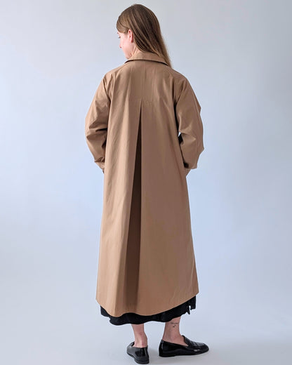 A woman is shown from behind wearing a long, straight camel-brown trench coat over a black skirt, standing against a plain light gray studio background.