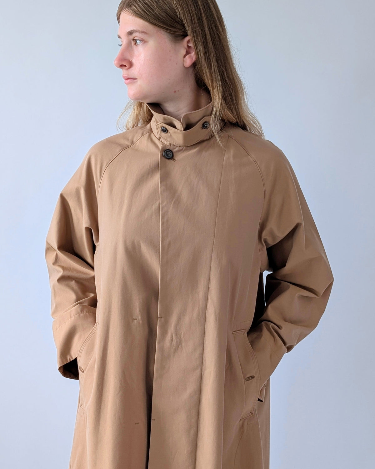 A woman wearing a long, loose-fitting tan coat with a high collar and dark buttons, photographed against a plain light gray studio background.